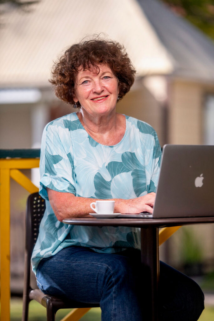 DSC05340 Copywriterin Bärbel Pfeiffer sitzt draußen am Café-Tisch mit Espresso und Laptop: lockiges Kurzhaar, gemustertes Oberteil, entspanntes Lächeln.